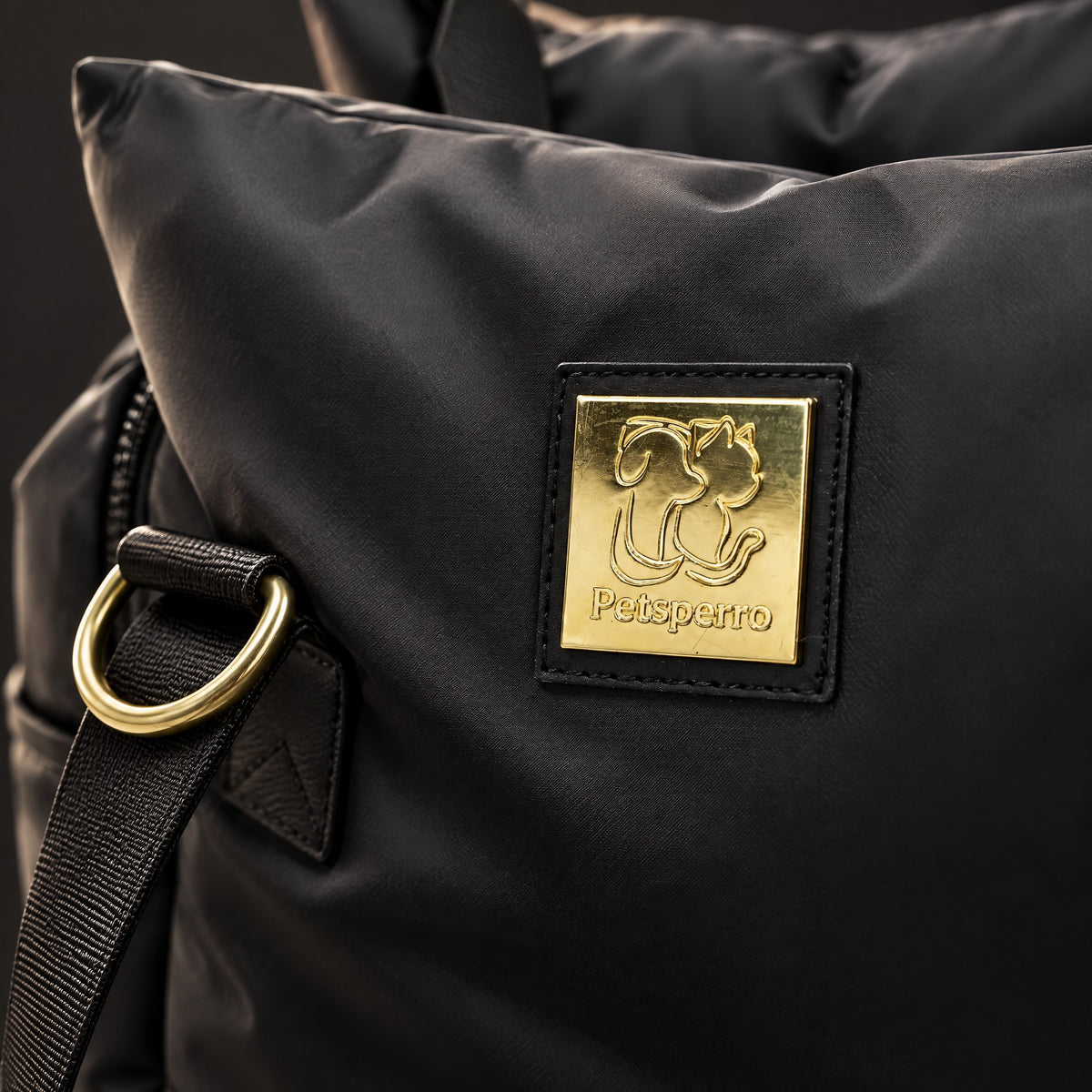 Macro close-up of the black Petsperro dog car seat: gold Petsperro nameplate on the pillow panel with a gold D-ring and strap
